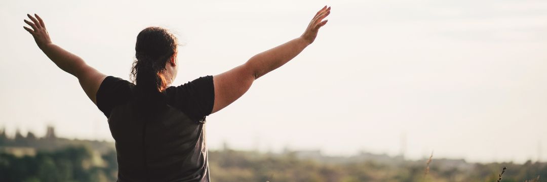 Obese woman looking out into field with arms up over her head